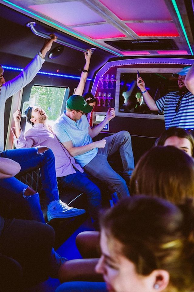 Friends dancing and laughing inside a neon-lit party bus with colorful LED ceiling lights and a small bar area.