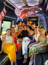 Group of friends smiling and playfully kissing one woman inside a decorated party bus with pink balloons, banner and LED lights.