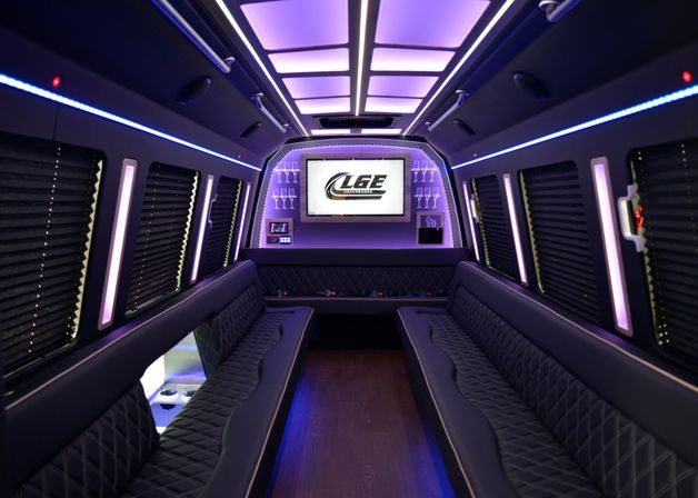 Luxury party bus interior with purple LED ambient lighting, opposing quilted leather bench seats, hardwood floor, built-in flat-screen TV and glass racks — modern limo shuttle vibe.