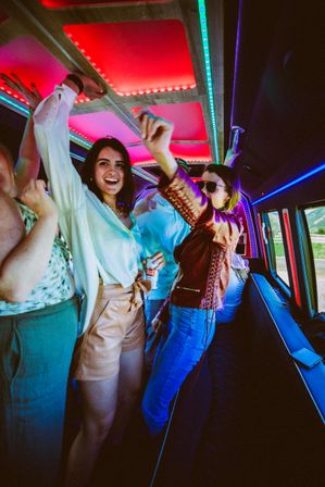 Group of friends dancing inside an LED-lit party bus with vibrant red and blue ceiling lights; smiling woman in a white blouse and leather shorts holds a drink as companions cheer near windows with countryside visible outside.
