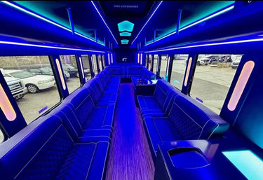 Vibrant blue-lit party bus interior with wraparound tufted leather benches, wood-plank aisle, overhead LED accents and windows showing parked cars outside
