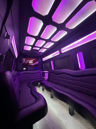 Party-ready bus interior with purple LED lighting, curved black leather bench seating, illuminated geometric ceiling panels, wood-look floor and front TV — luxury group transport.