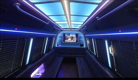 LED-lit luxury party bus interior with blue ceiling panels, opposing quilted leather bench seats, window blinds, wall-mounted TV displaying an aquarium, built-in cup holders and ambient party lighting