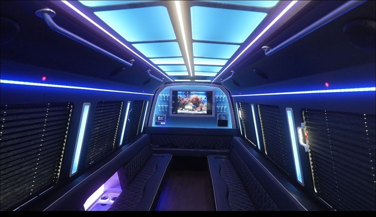 LED-lit luxury party bus interior with blue ceiling panels, opposing quilted leather bench seats, window blinds, wall-mounted TV displaying an aquarium, built-in cup holders and ambient party lighting