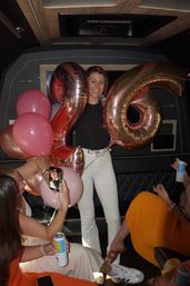 Smiling woman posing with large rose-gold "26" balloons inside a party bus, friends snapping photos and holding drinks for a fun birthday celebration