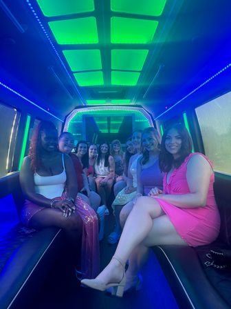 Group of smiling women in colorful party dresses seated inside a neon-lit party bus with green ceiling panels and blue LED trim, posing for a night-out celebration.