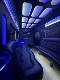 Sleek luxury party bus interior with curved black leather bench seating, wood-look floor, window blinds, and blue LED ceiling panels and ambient accent lighting