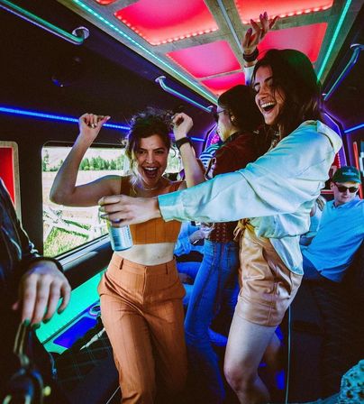 Friends dancing and laughing inside a neon-lit party bus with colorful LED ceiling panels, one woman holding a canned drink and a countryside view visible through the window.
