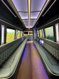 LED-lit luxury party van interior with long quilted black leather benches, wood floor, and panoramic windows revealing green fields and distant mountains.
