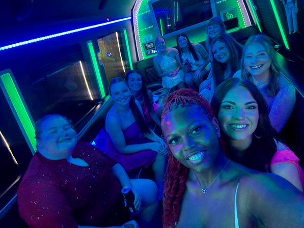 Group selfie of women in dresses inside a neon-lit party bus or stretch limo, smiling under green and blue LED lights for a nighttime celebration.
