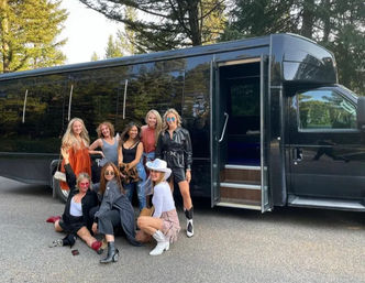 Group of eight women in stylish outfits posing by a black party bus parked on a tree-lined road, some standing and some crouching, ready for a road trip.