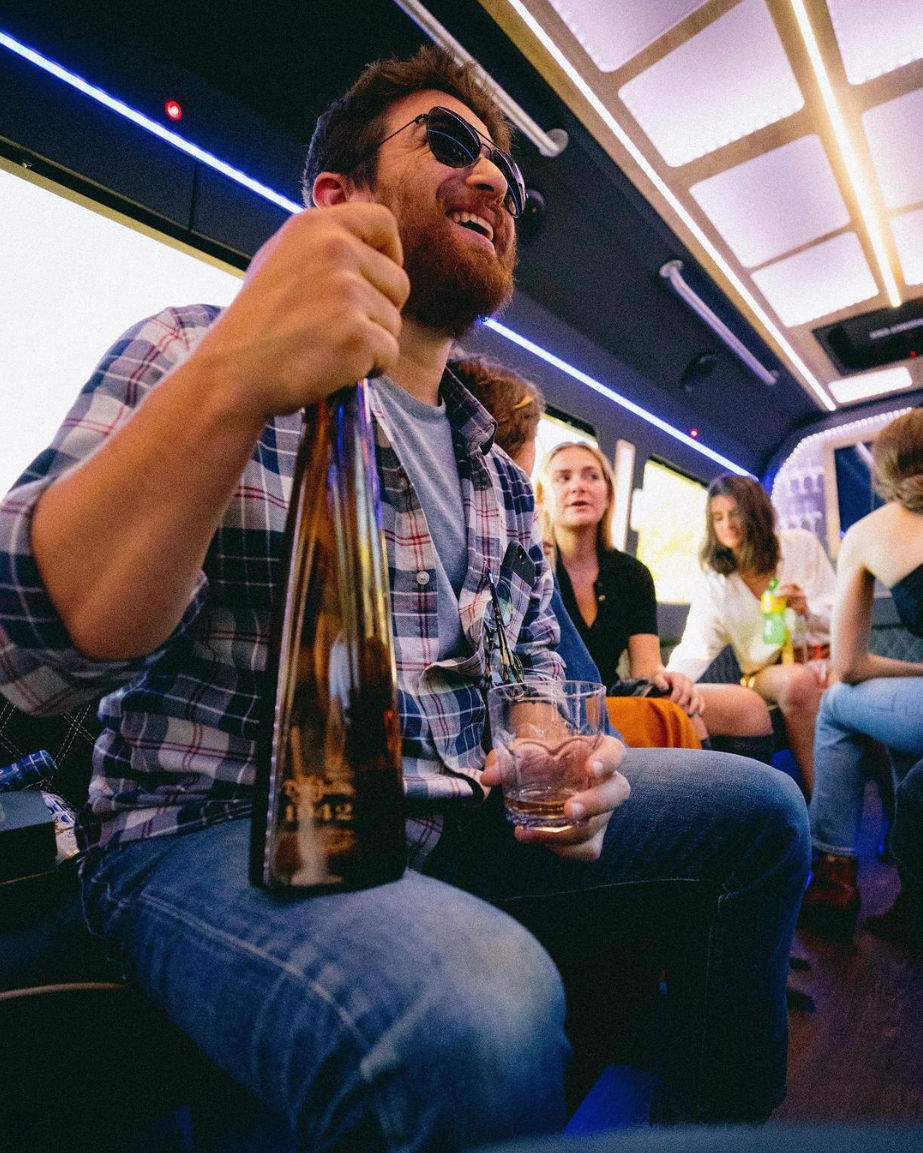 Bearded man in sunglasses laughing on a neon-lit party bus, holding a large bottle and a glass while friends chat