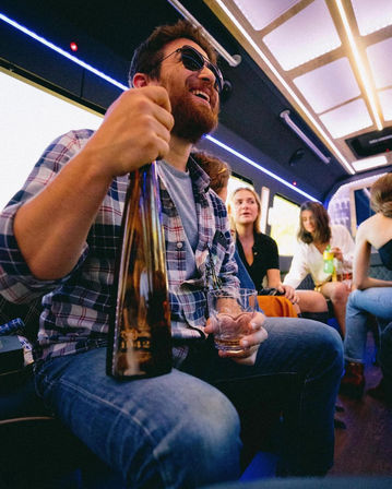 Bearded man in sunglasses laughing on a neon-lit party bus, holding a large bottle and a glass while friends chat