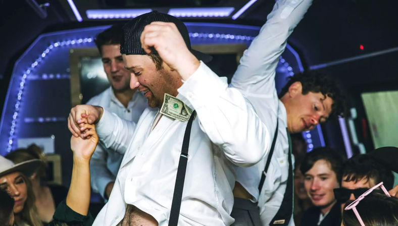 Friends partying inside a lit party bus, a man in a white shirt and suspenders dances with a dollar tucked into his shirt while others cheer.