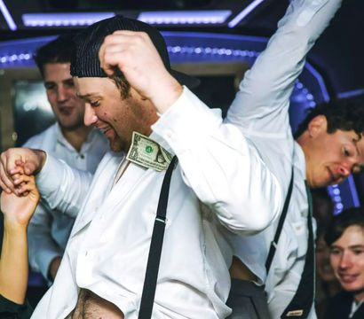 Friends partying inside a lit party bus, a man in a white shirt and suspenders dances with a dollar tucked into his shirt while others cheer.