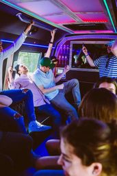 Group of friends partying inside a neon-lit party bus interior with colorful LED ceiling lights, people laughing, dancing on bench seats and recording videos on their phones.