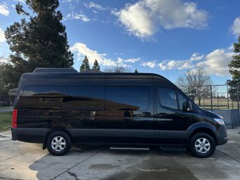 Sleek black high-roof camper van parked on a concrete driveway near a neighborhood park and basketball court, framed by evergreen trees under a bright blue sky with scattered clouds.