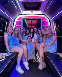 Group of ten women in blue dresses smiling and posing inside a neon-lit party bus with colorful LED lights and leather bench seating, celebrating a night out