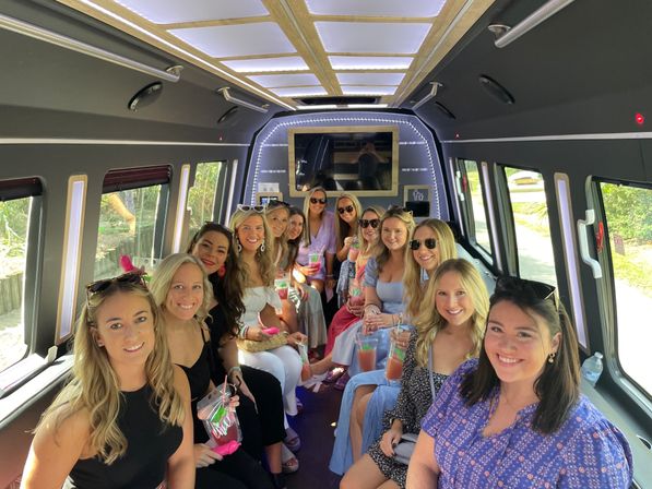 Cheerful group of women holding colorful drinks inside a lighted party bus, seated along both sides with sunny greenery visible through the windows.
