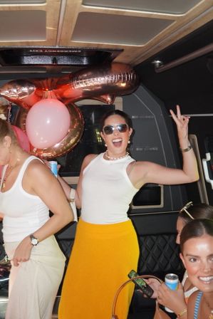 Smiling women celebrating inside a party bus with rose-gold and pink balloons; one woman in sunglasses, a white tank top and bright yellow skirt flashes a peace sign.