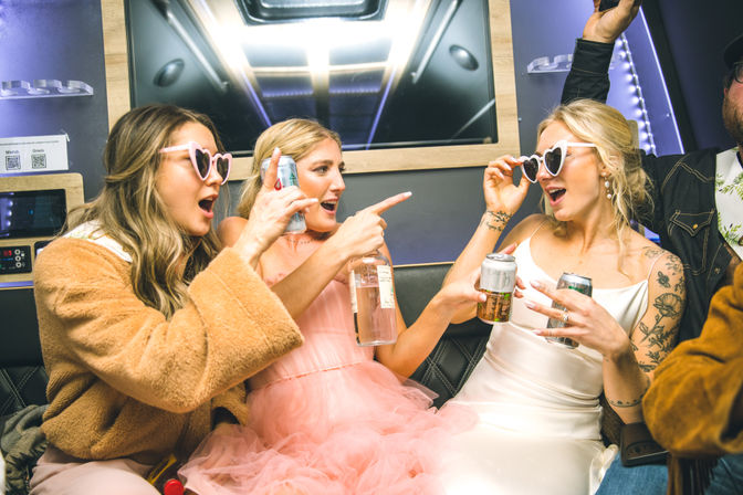 Bachelorette celebration inside a lit party bus: three women — bride in a white dress, friend in a pink tulle dress, and a woman in a cozy coat — wearing heart-shaped sunglasses, holding drinks, laughing and pointing.