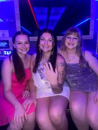 Three smiling friends in sparkly party dresses inside a neon-lit party bus; center wearing a bachelorette sash and showing an engagement ring with visible arm tattoos.