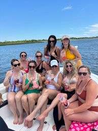 Nine women in colorful swimsuits smiling and holding fruity cocktails with curly pink straws while seated on a boat in a calm coastal inlet under a clear blue sky — sunny summer boating celebration.