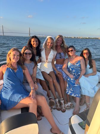 Seven friends on a boat at sunset, bride-to-be in a white romper and veil in the center, all smiling in summer dresses and sandals with a cable-stayed bridge and waterfront skyline in the background.