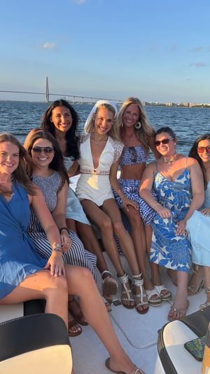 Seven friends on a boat at sunset, bride-to-be in a white romper and veil in the center, all smiling in summer dresses and sandals with a cable-stayed bridge and waterfront skyline in the background.