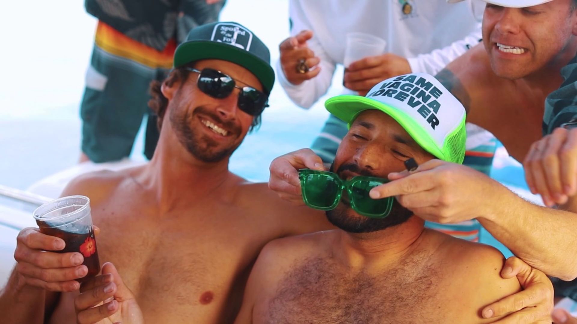 Shirtless friends laughing on a sunny boat party at sea, wearing trucker hats and sunglasses as one holds a drink and another playfully fits oversized green novelty glasses on a friend.