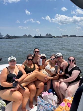 Seven friends in swimsuits smiling and cuddled together on a boat during a sunny summer harbor outing, with calm water and large navy ships docked on the distant shoreline.