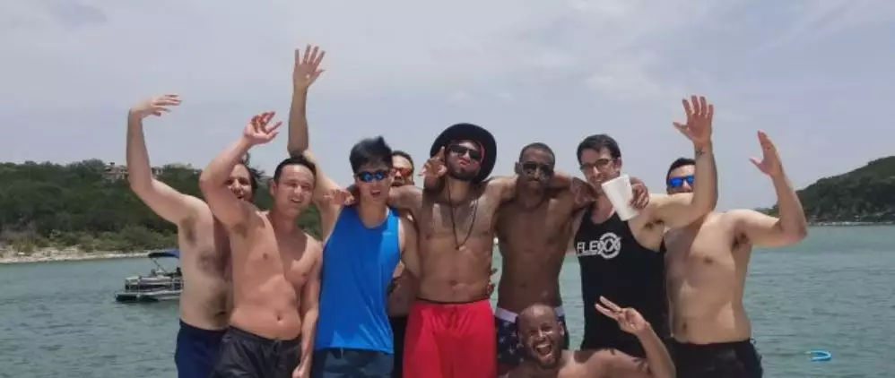 Cheerful group of friends posing and waving on a boat during a sunny summer lake day with a tree-lined shoreline in the background