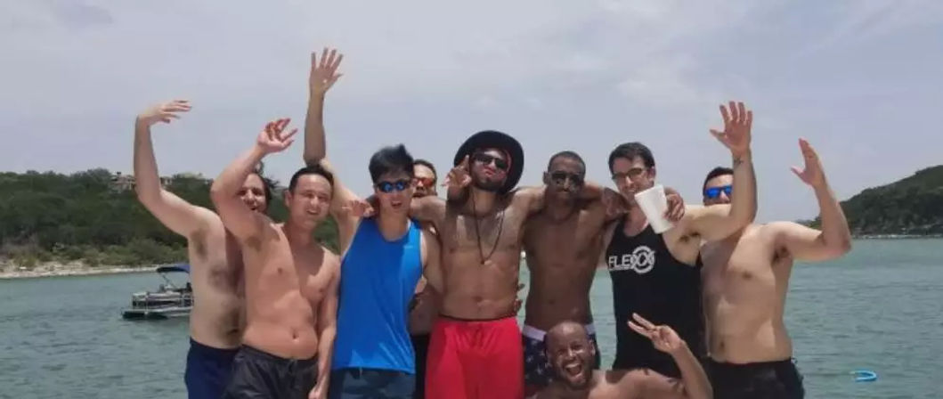 Cheerful group of friends posing and waving on a boat during a sunny summer lake day with a tree-lined shoreline in the background