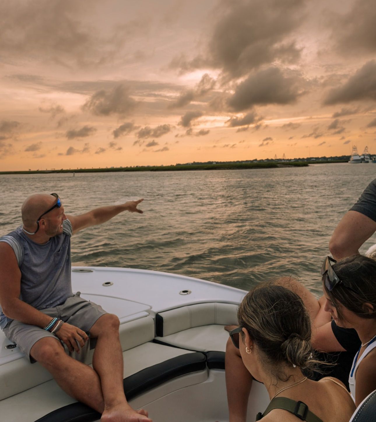 Friends relaxing on a boat bow at sunset, a man pointing toward a marshy coastal inlet under dramatic orange-gray clouds and calm water
