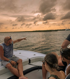 Friends relaxing on a boat bow at sunset, a man pointing toward a marshy coastal inlet under dramatic orange-gray clouds and calm water