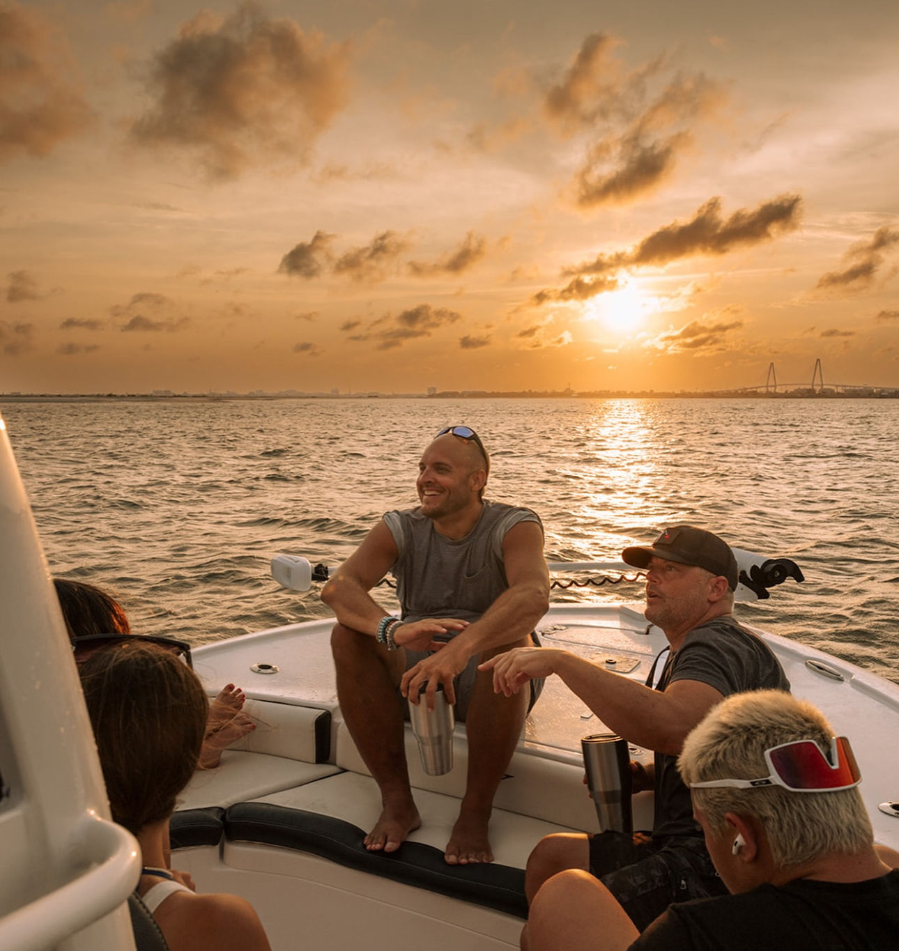 Sunset boat cruise on calm coastal waters — a group relaxing on a motorboat, a smiling man holding a stainless tumbler, golden sky and sun reflecting on the water with a distant suspension bridge silhouette.