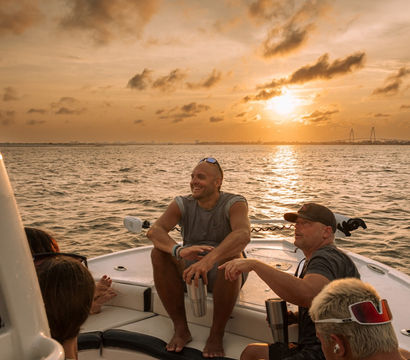 Sunset boat cruise on calm coastal waters — a group relaxing on a motorboat, a smiling man holding a stainless tumbler, golden sky and sun reflecting on the water with a distant suspension bridge silhouette.