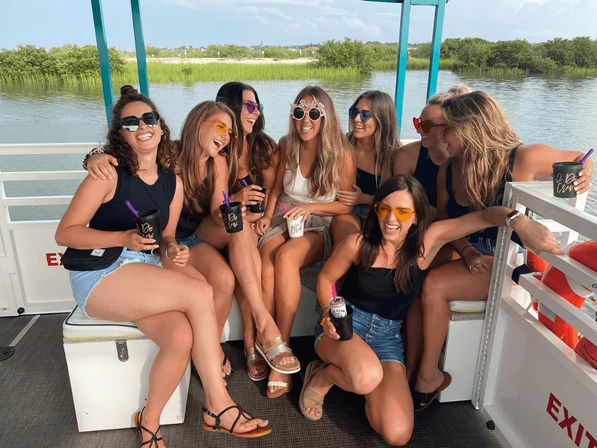 Eight friends in sunglasses and summer outfits laughing on a pontoon boat over marshy coastal water, holding drinks and enjoying a sunny waterfront outing.