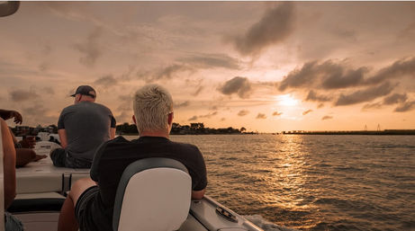 Passengers on a boat enjoying a golden sunset over coastal waters, silhouetted against a glowing sky with a distant shoreline and bridge
