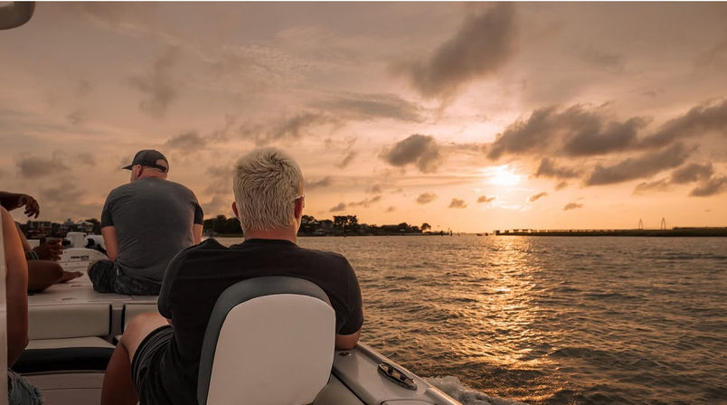 Passengers on a boat enjoying a golden sunset over coastal waters, silhouetted against a glowing sky with a distant shoreline and bridge