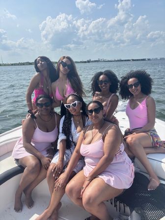 Seven women in pink swimsuits posing on a motorboat during a sunny summer cruise on calm bay waters under a blue sky with fluffy clouds