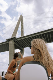 Two women on a boat looking up at a towering cable-stayed bridge with concrete pylons and radiating cables against a partly cloudy sky