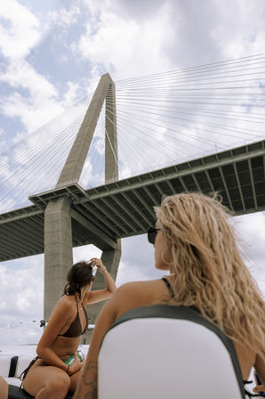 Two women on a boat looking up at a towering cable-stayed bridge with concrete pylons and radiating cables against a partly cloudy sky