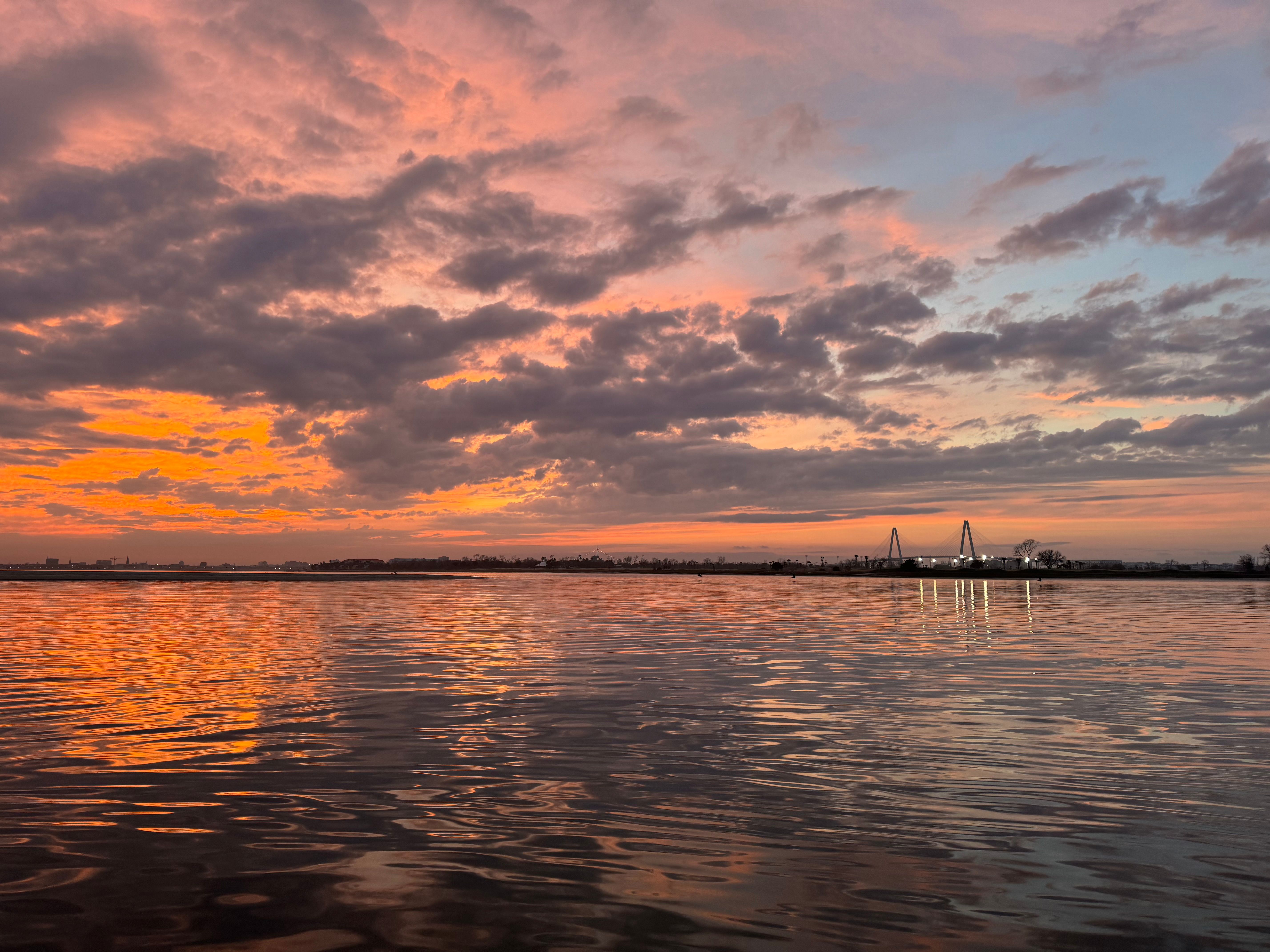 Pink-orange sunset over a calm waterfront with dramatic clouds, rippling reflections and a distant lit cable-stayed bridge on the horizon