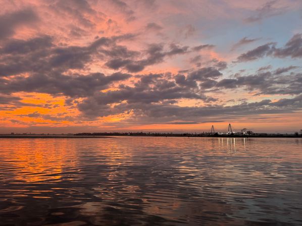 Pink-orange sunset over a calm waterfront with dramatic clouds, rippling reflections and a distant lit cable-stayed bridge on the horizon