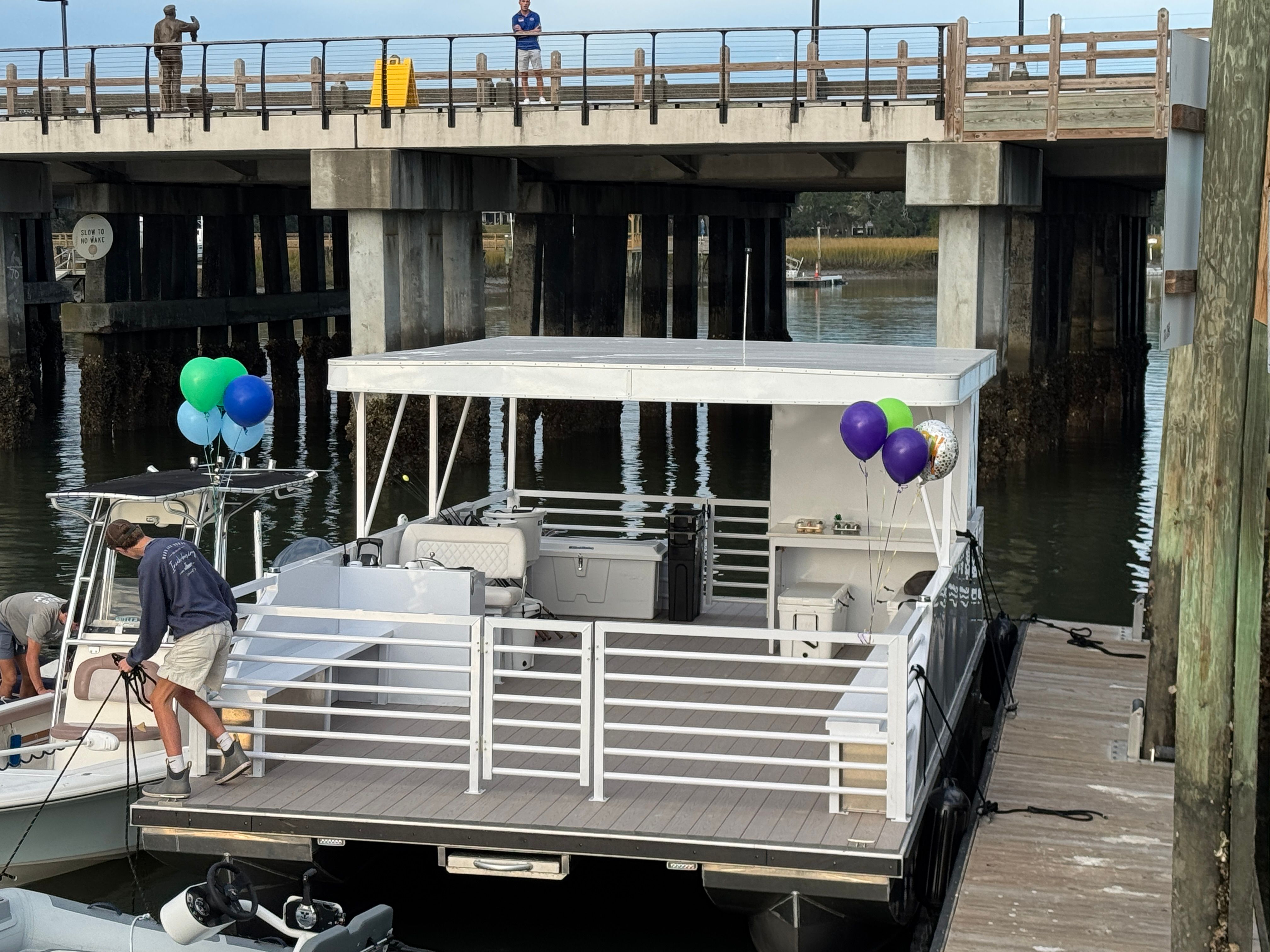 Party-ready white pontoon boat with blue, green, and purple balloons moored at a marina dock beneath a concrete bridge, a person securing ropes on the bow.