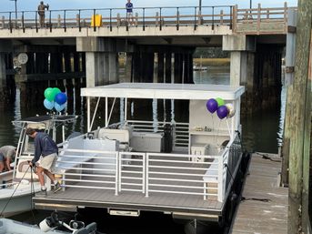 Party-ready white pontoon boat with blue, green, and purple balloons moored at a marina dock beneath a concrete bridge, a person securing ropes on the bow.