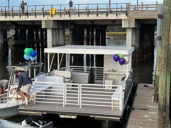 Party-ready white pontoon boat with blue, green, and purple balloons moored at a marina dock beneath a concrete bridge, a person securing ropes on the bow.
