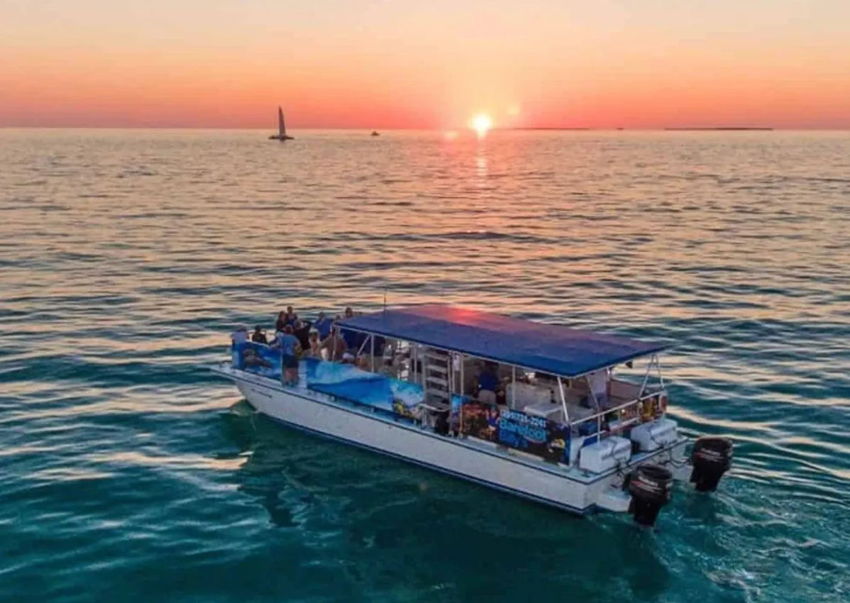 Sunset cruise on calm turquoise ocean — covered tour boat with passengers gliding toward a glowing orange horizon, small sailboat visible in the distance.