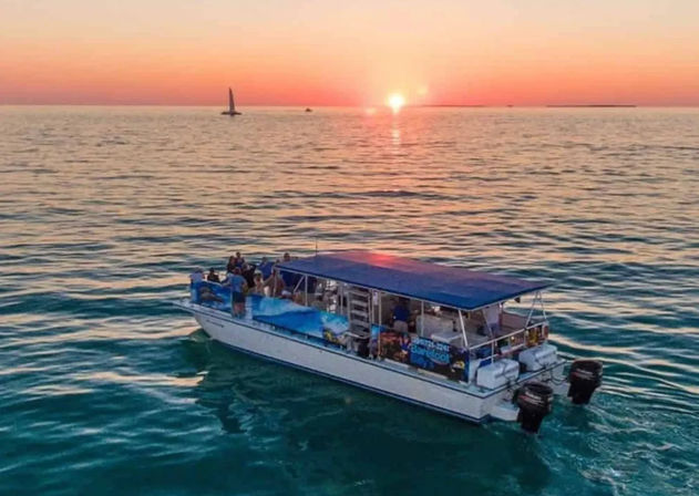 Sunset cruise on calm turquoise ocean — covered tour boat with passengers gliding toward a glowing orange horizon, small sailboat visible in the distance.